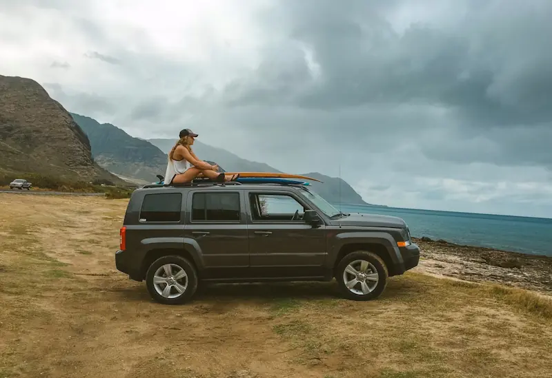 Woman sitting on an SUV with surfboards on the roof parked by a scenic coastal road