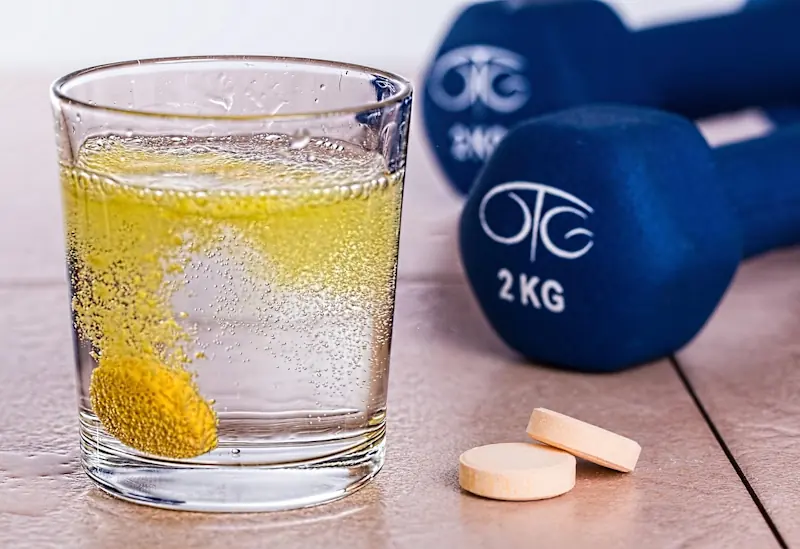 Glass of water with electrolyte tablet fizzing and two dumbbells on a tiled floor