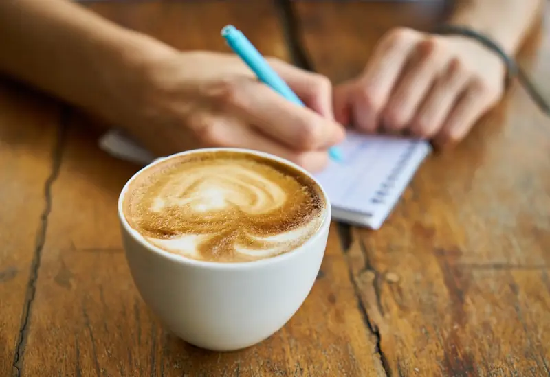 Person sitting at a table with a coffee and writing in a notepad