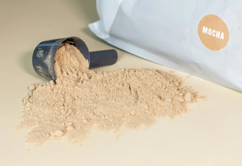 A scoop of mocha whey protein powder poured onto a surface next to its packaging
