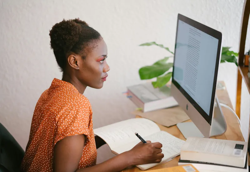 Black woman writing in a notebook at a desk with an iMac and books nearby
