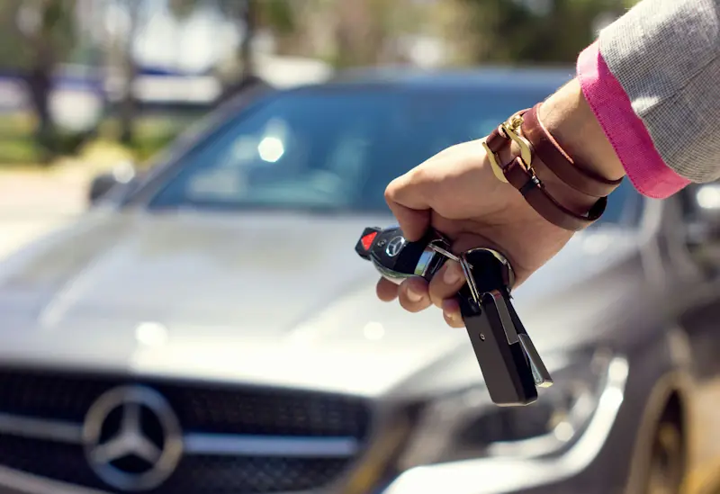 Hand holding car keys in front of a parked car, representing everyday car ownership costs.