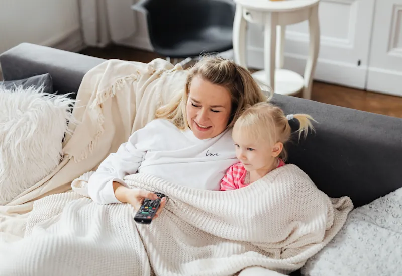 Adult and child sitting on a sofa wrapped in a knitted blanket while watching TV.