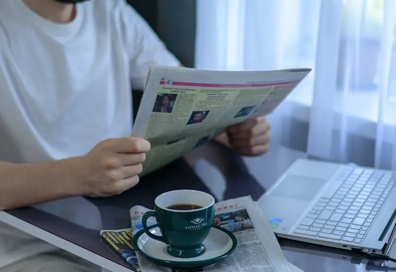 Person reading newspapers with a cup of tea and a laptop on the table.