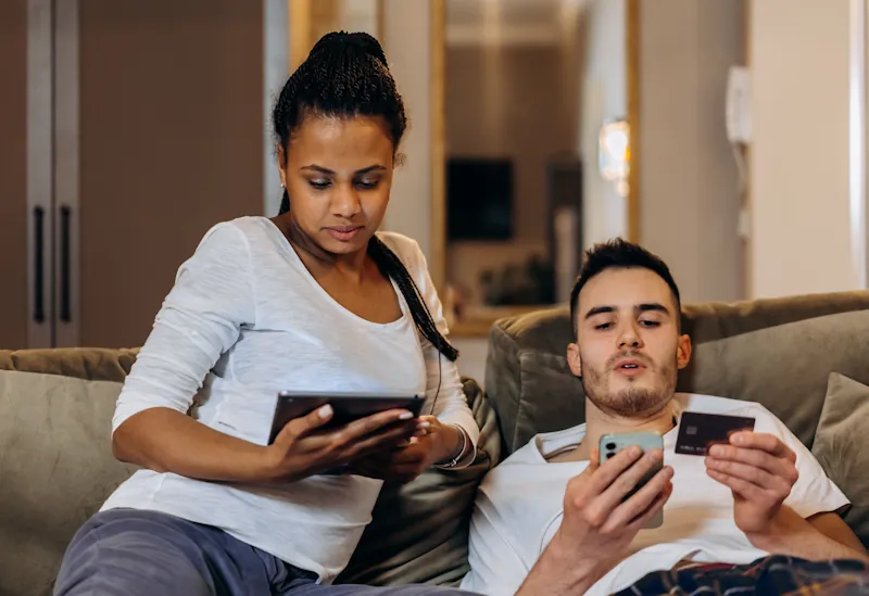 Person sitting on a sofa holding a credit card and smartphone while online shopping.