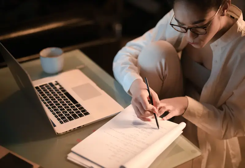 A person writing on papers at a desk while working on a laptop