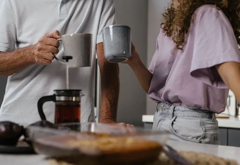 Two people standing in a kitchen holding mugs with food on the counter, showing everyday food and drink habits.