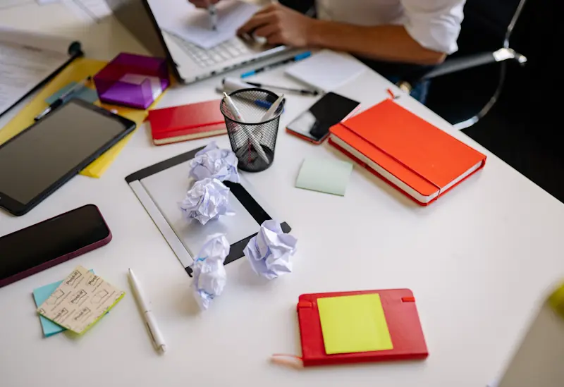 A cluttered desk with a laptop, phone, tablet, sticky notes, notebooks, and crumpled papers