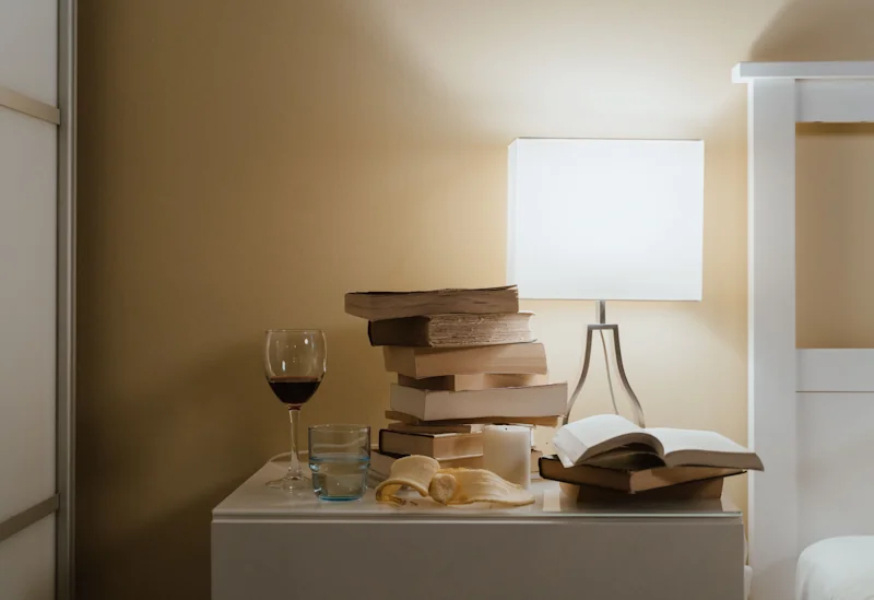 A stack of old books and an open book beside a lit table lamp on a bedside table