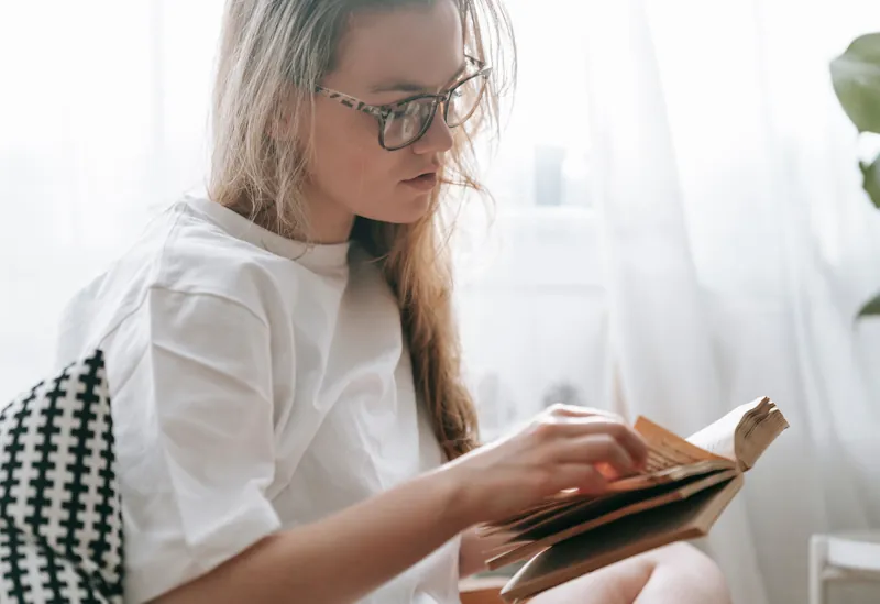 A person reading an open book with aged pages near a window in soft light