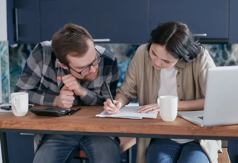 Two people reviewing household bills with a calculator and laptop in a kitchen, representing the hidden costs of owning a home in the UK.