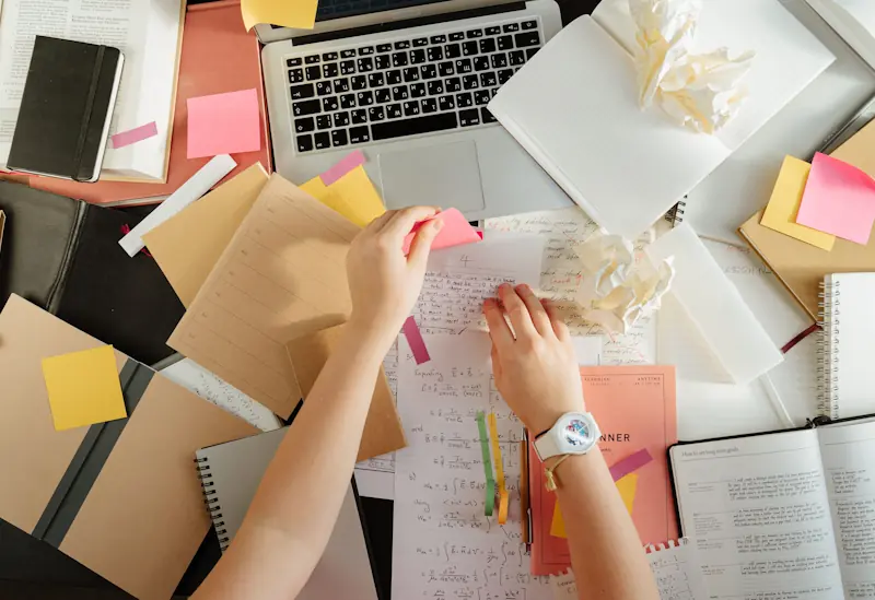 A cluttered desk with a laptop, books, planners, sticky notes, and crumpled papers while someone organises notes