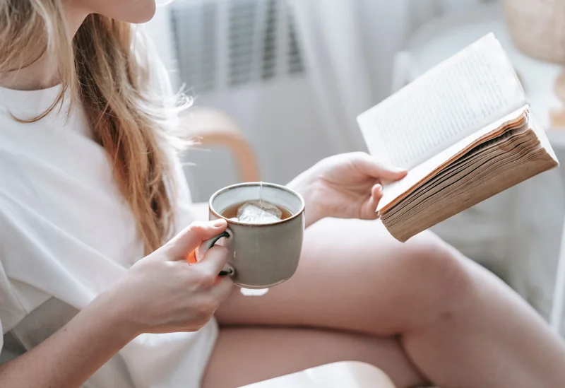 A person holding a mug while reading an open book in soft natural light