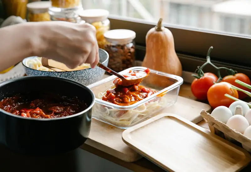 A person spooning pasta sauce into a glass meal prep container in a home kitchen