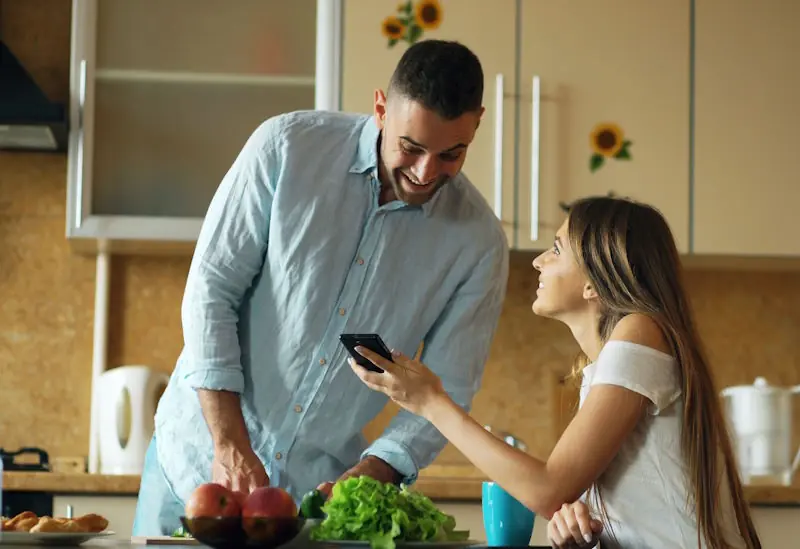 Two people in a kitchen checking something on a phone while sorting out household tasks