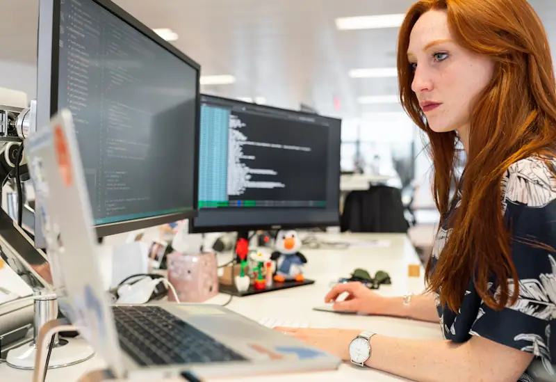 A person using a laptop and dual monitors with several windows open on a busy desk
