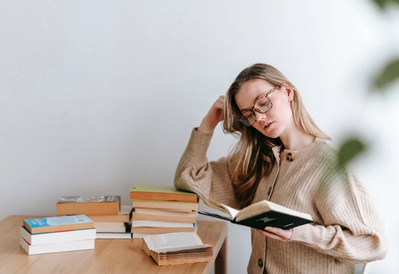 A person in glasses reading at a wooden table with multiple stacks of books in a calm indoor setting