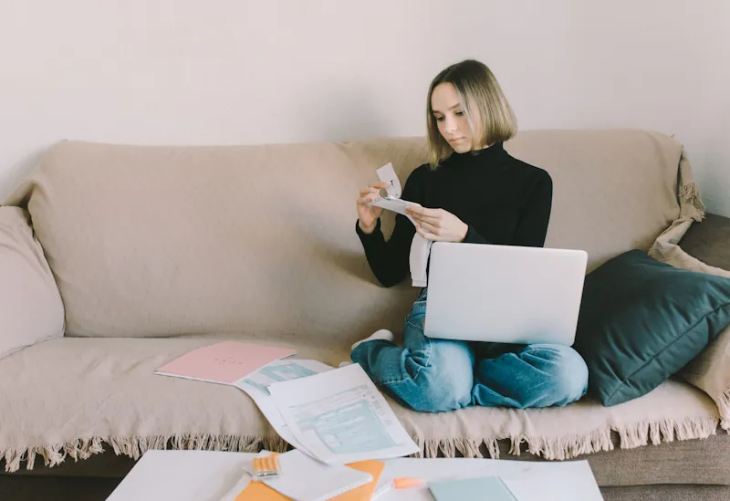 A person sitting on a couch with a laptop and scattered papers, looking over receipts