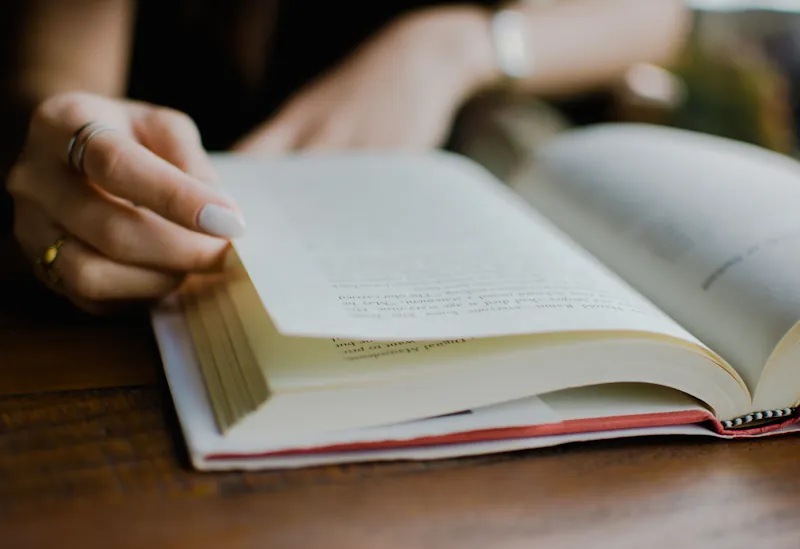 A hand turning a page in an open red‑covered book on a wooden table