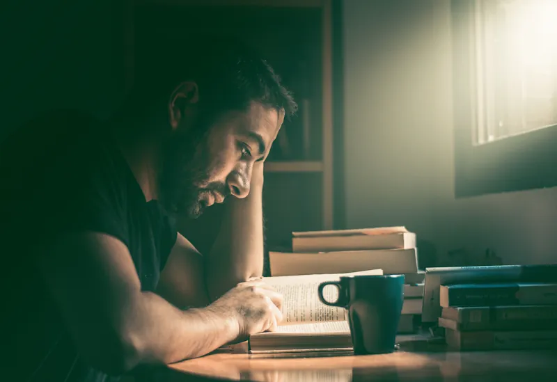 A person reading a large classic novel at a wooden table with a stack of books and a mug nearby