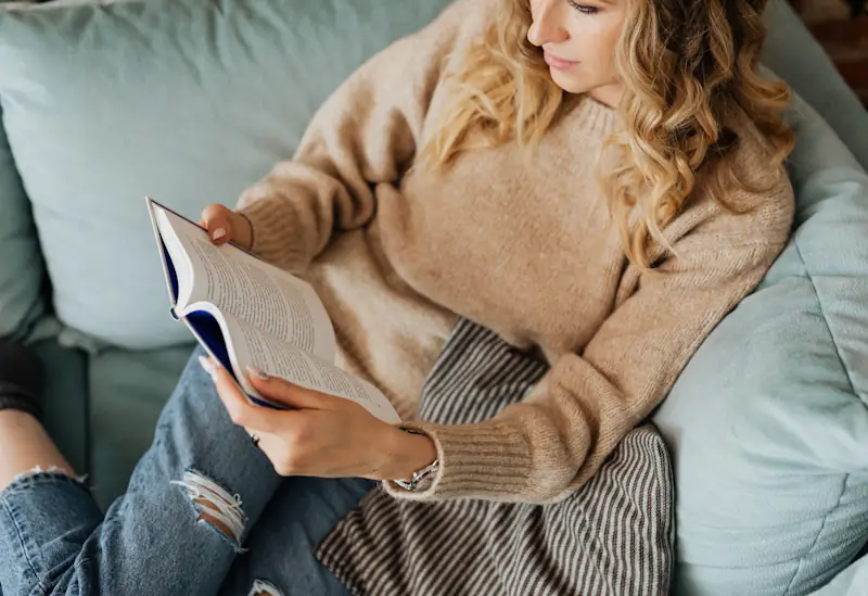 A person sitting on a couch reading an open book in soft natural light