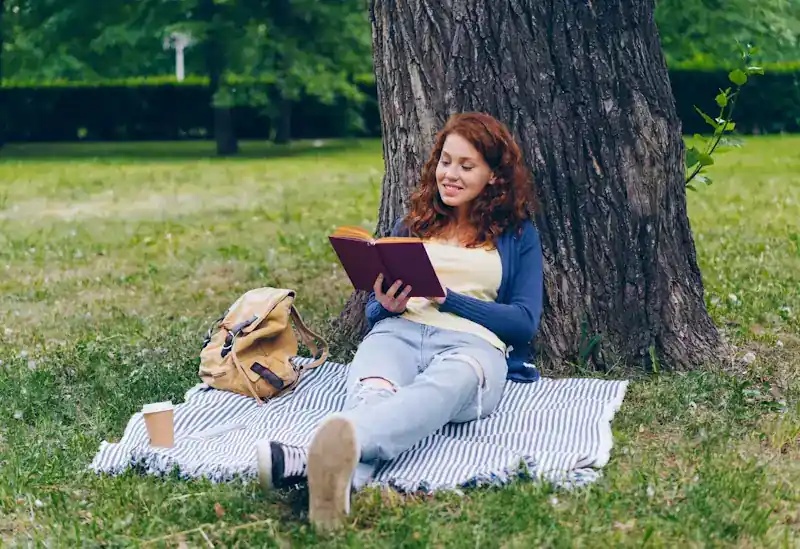 A person sitting under a tree reading a paperback book in natural light