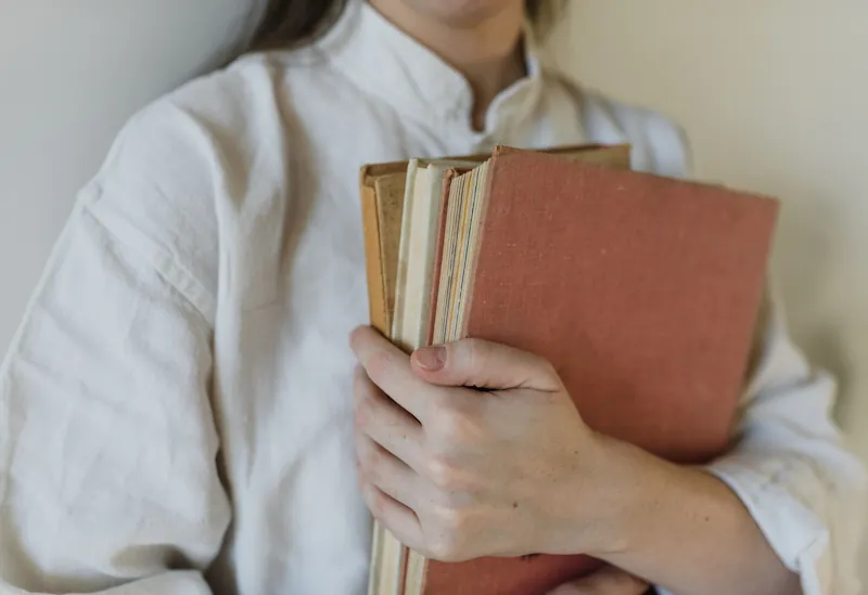 A person holding a small stack of classic hardback books in soft natural light
