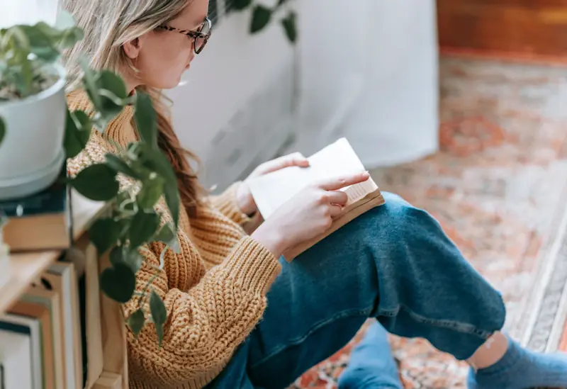 A young person reading a paperback book beside a bookshelf in natural light