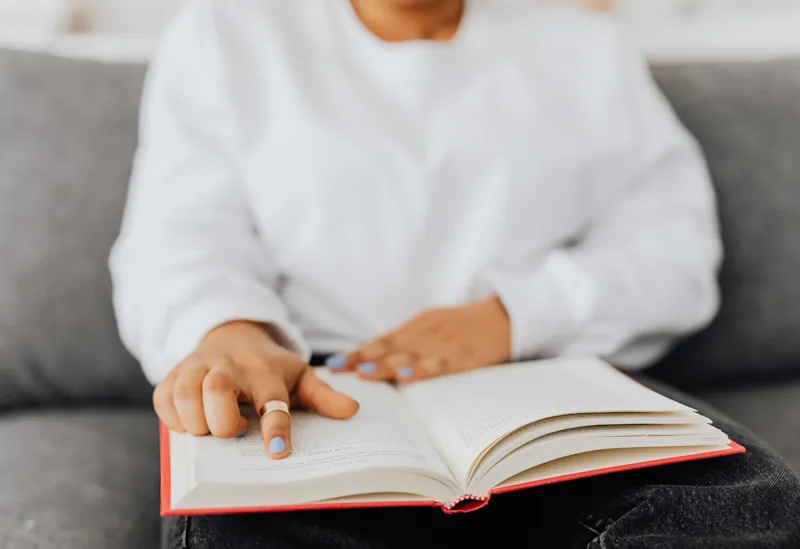 A person sitting on a couch reading an open book with a red cover