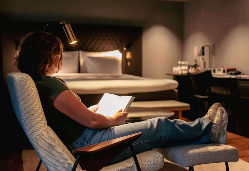 A person reading a large book in a cosy armchair under warm lamp light