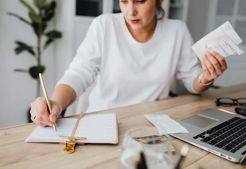 Person reviewing receipts and writing in a notebook at a desk with a laptop and phone, representing how everyday living costs add up.