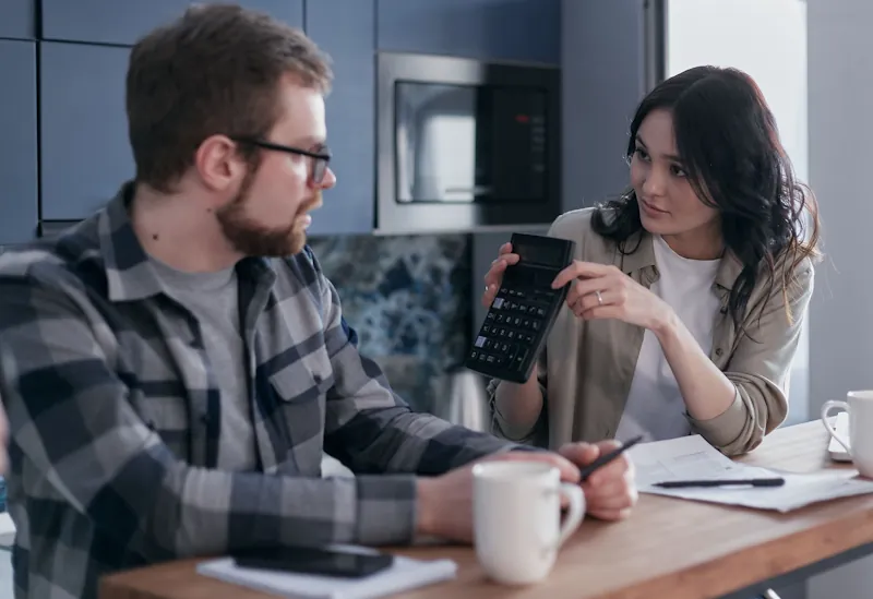 Two adults reviewing household expenses at a kitchen table with a calculator, papers and a laptop, representing the real cost of raising a family in the UK.