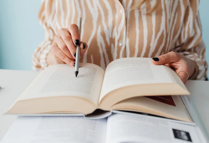 A person sitting at a desk reading an open book, pointing at a line of text with a pen, with another book beneath it.