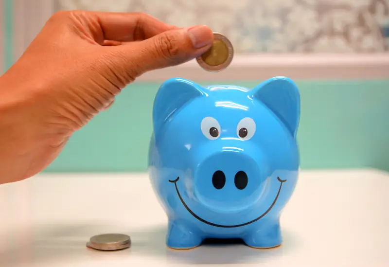 A hand holding a coin above a blue piggy bank on a table