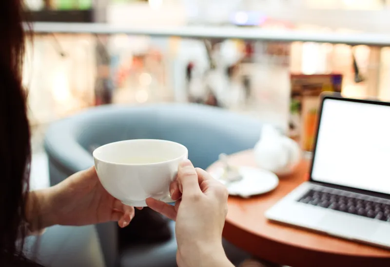 Person holding a cup of coffee at a café table, representing small daily spending habits that add up over a year..