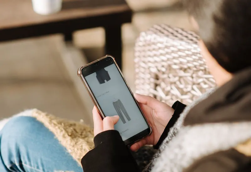 Person browsing a clothing item on a smartphone while holding a drink, representing smart shopping choices that help you spend less and get more.