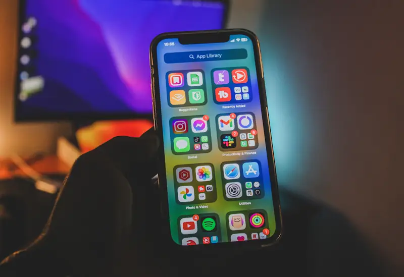 A smartphone displaying grouped digital apps such as social media, productivity and finance, with a blurred computer screen behind it, illustrating everyday digital habits.