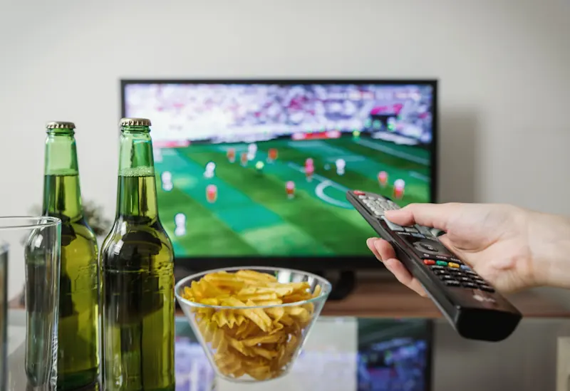 A person holding a remote while watching a football match with snacks on the table