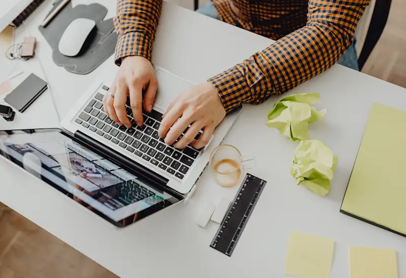A cluttered desk with a laptop, phone, sticky notes, crumpled papers, and scattered stationery