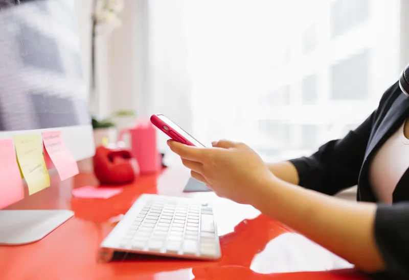 A person checking their phone at a cluttered desk with sticky notes and work tasks visible